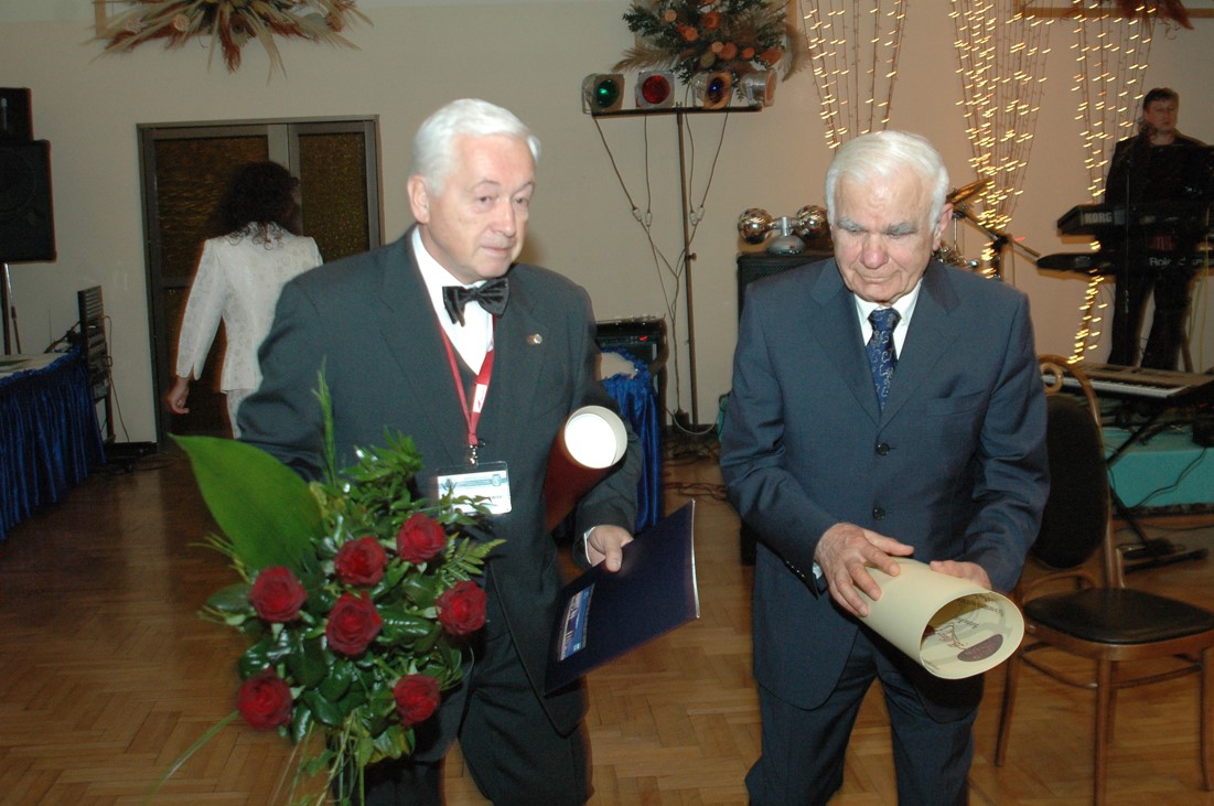 Na pierwszym planie zdjęcia widać dw&oacute;ch mężczyzn o siwych włosach, kt&oacute;rzy są ubrani w garnitury. Mężczyzna po lewej stronie trzyma bukiet kwiat&oacute;w, tubę na dokumenty lub plakaty i teczkę, mężczyzna po prawej trzyma zwinięty w rulon dokument lub plakat. Za nimi widać innych ludzi i światełka ozdobne